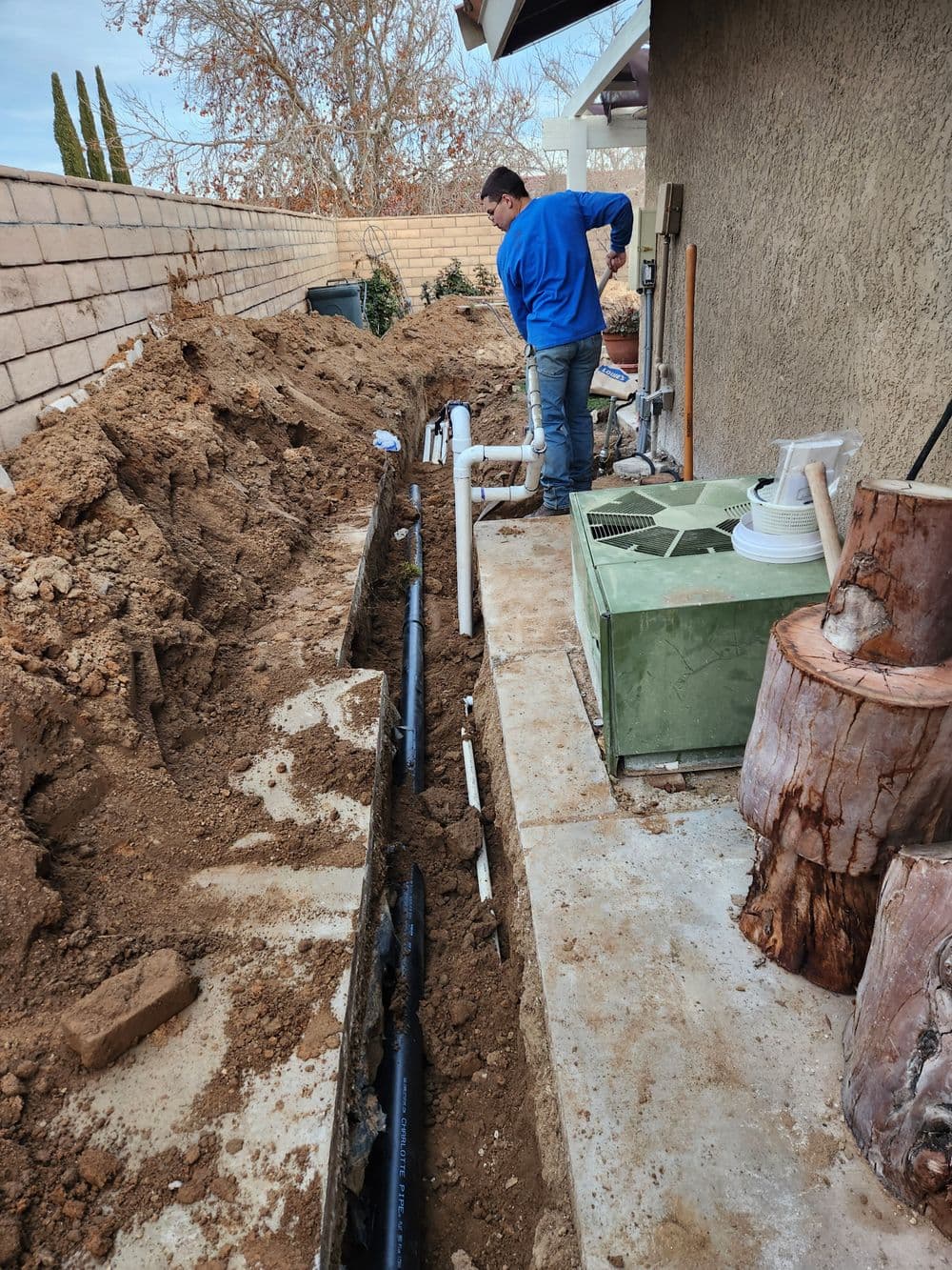 Worker installing plumbing in a trench outside a home, surrounded by dirt and equipment.