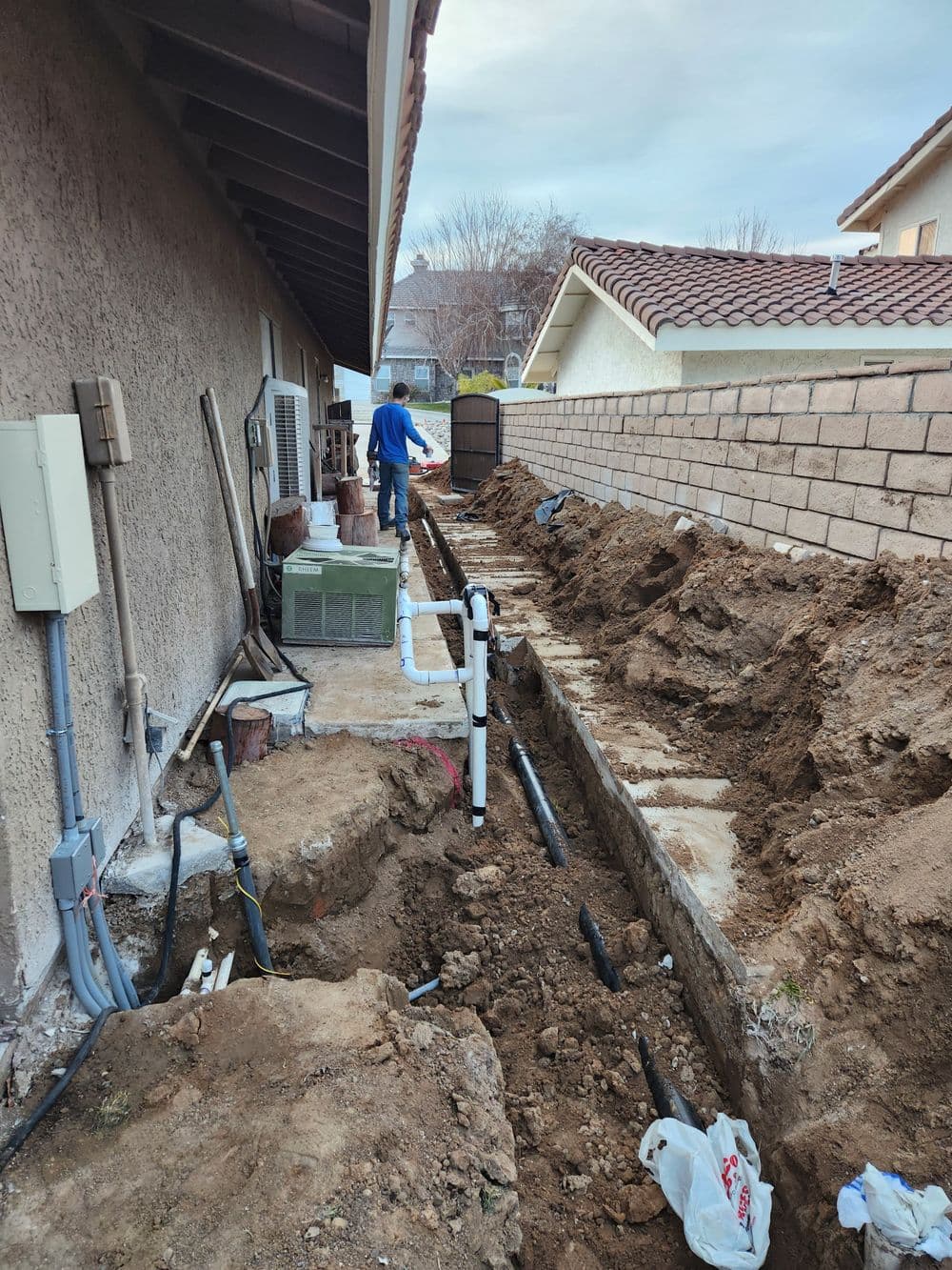 Workman installing plumbing in a trench beside a house, with exposed pipes and dirt.